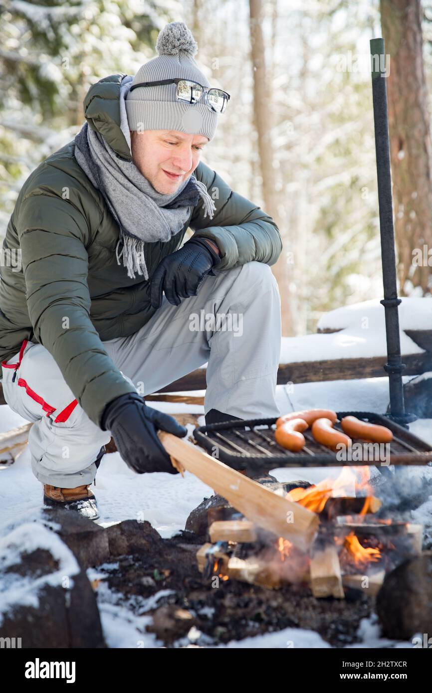 Man roasting sausages on campfire in forest by the lake, making a fire, grilling. Happy tourist exploring Finland. Beautiful sunny winter landscape Stock Photo