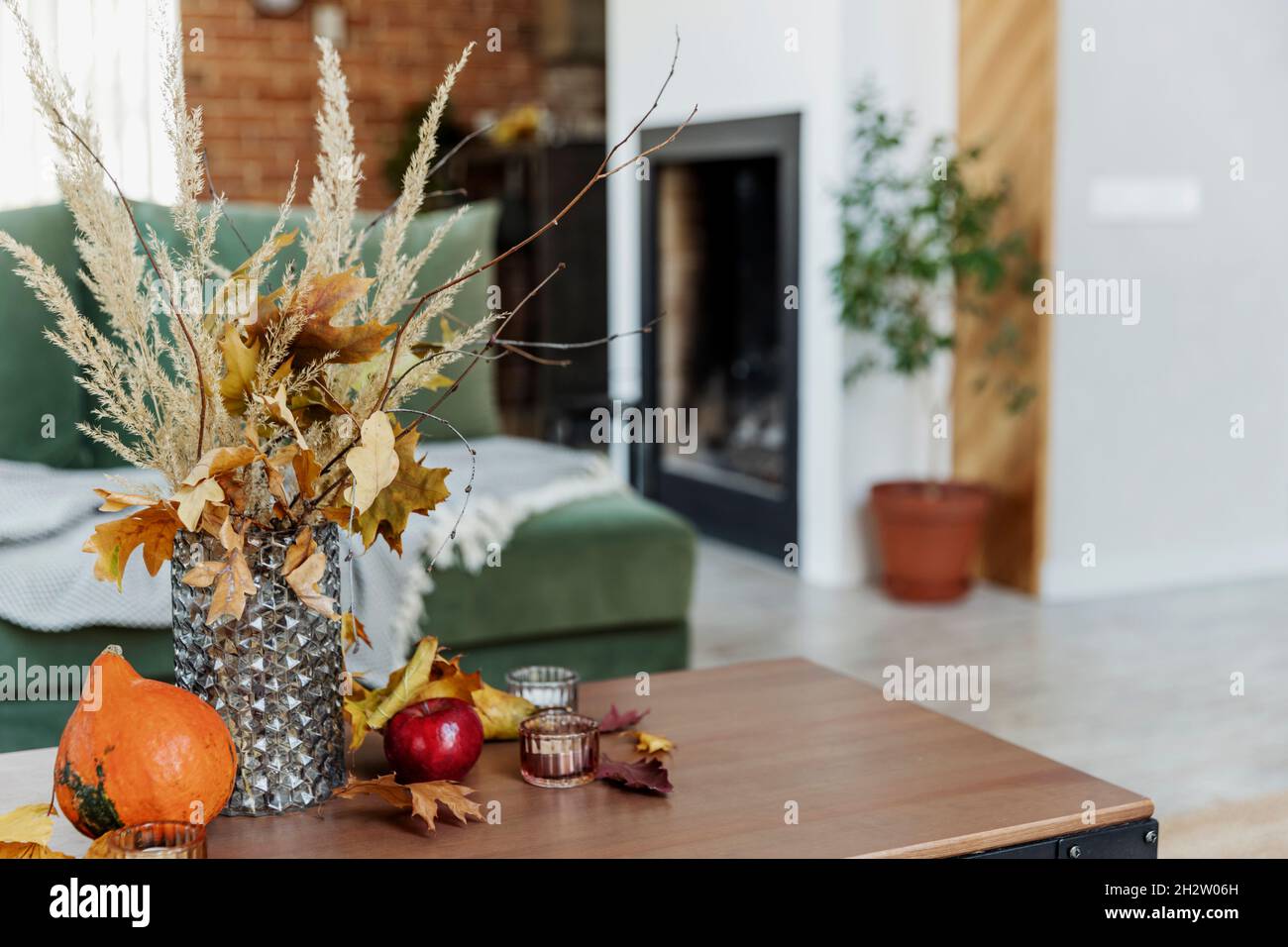 Autumn time decorated home interior. Yellow flowers bouquet in glass vase on wooden stylish table, pumpkin, red apples, leaves, candles, blanket. Gree Stock Photo