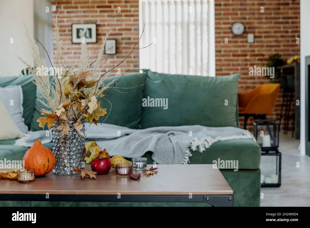 Autumn time decorated home interior. Yellow flowers bouquet in glass vase on wooden stylish table, pumpkin, red apples, leaves, candles, blanket. Gree Stock Photo