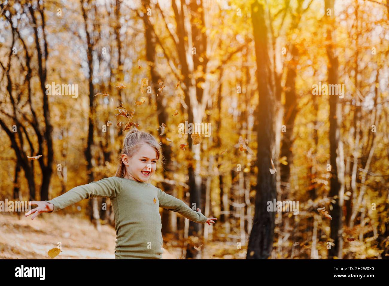 Small happy smiling little caucasian child girl playing with yellow leaves in autumn forest or garden around trees. Kid throws leaves up and they are Stock Photo