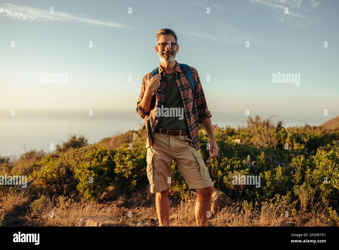 Male hiker looking at the camera on a hilltop. Happy mature hiker smiling while standing on top of a hill with a backpack. Adventurous male backpacker Stock Photo