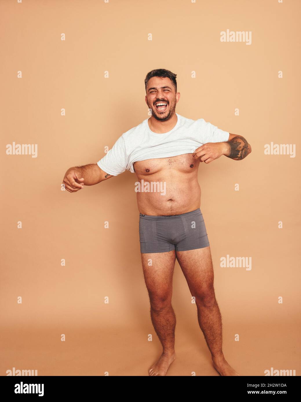 Flexing my natural body. Body positive young man lifting his shirt and smiling cheerfully while standing in a studio. Self-assured young man feeling c Stock Photo