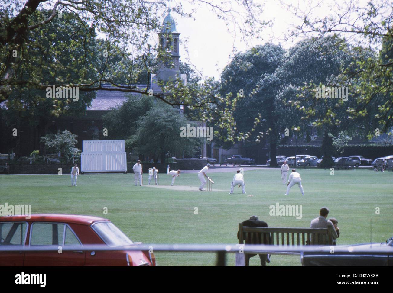 A cricket match involving members of the Kew Cricket Club, at Kew Green, west London in the early 1970s. Spectators are watching the match from a park bench and St Anne's Church lies beyond the cricket ground. Stock Photo