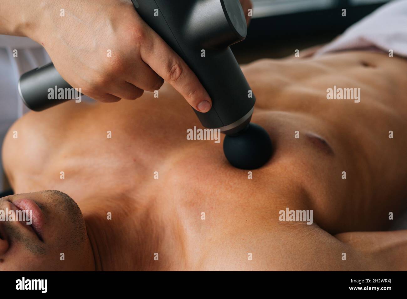 Close-up of unrecognizable professional male masseur receiving chest muscles using massage gun percussion tool of muscular athlete man Stock Photo