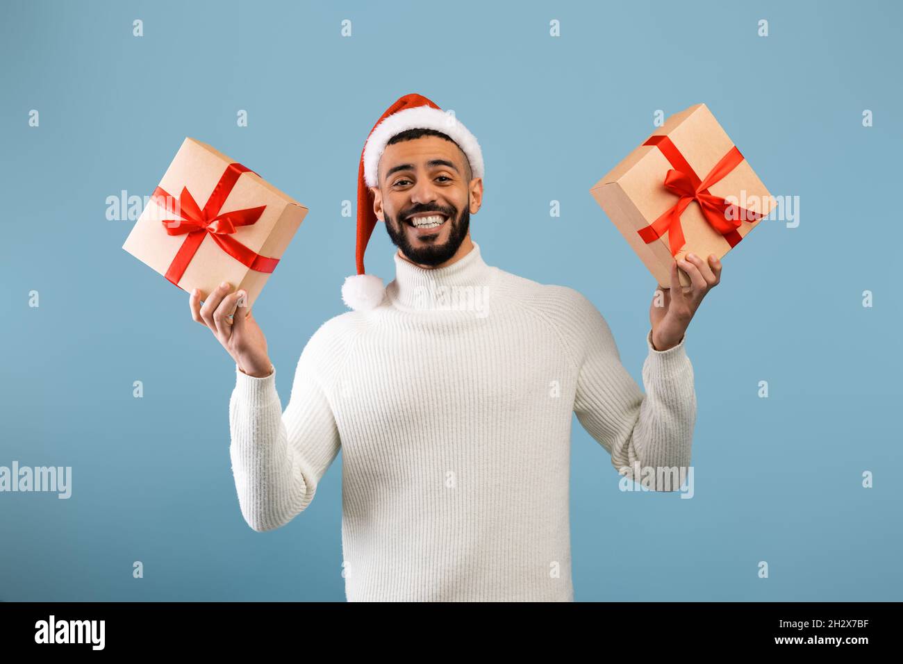 Happy arab guy in santa hat holding christmas or new year gifts and smiling at camera, standing over blue background Stock Photo