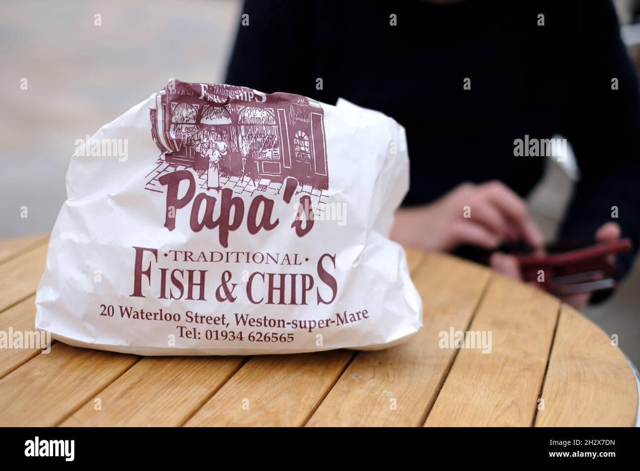 A paper bag of traditional english fish and chips placed on an outdoor table waiting to be opened Stock Photo
