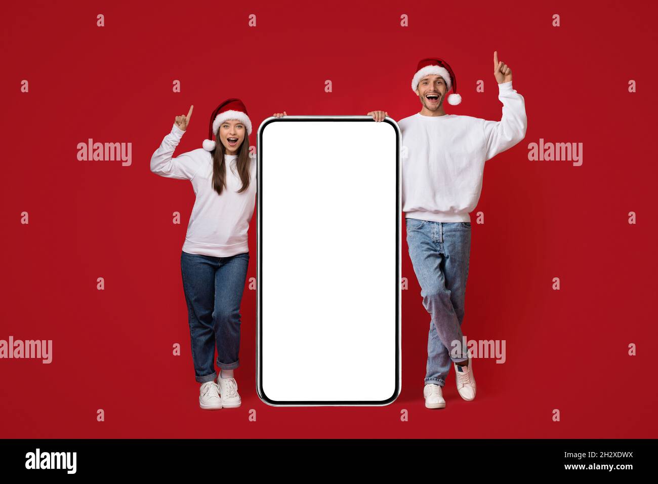 Chistmas Idea Concept. Couple In Santa Hats Standing With Big Blank Smartphone Stock Photo
