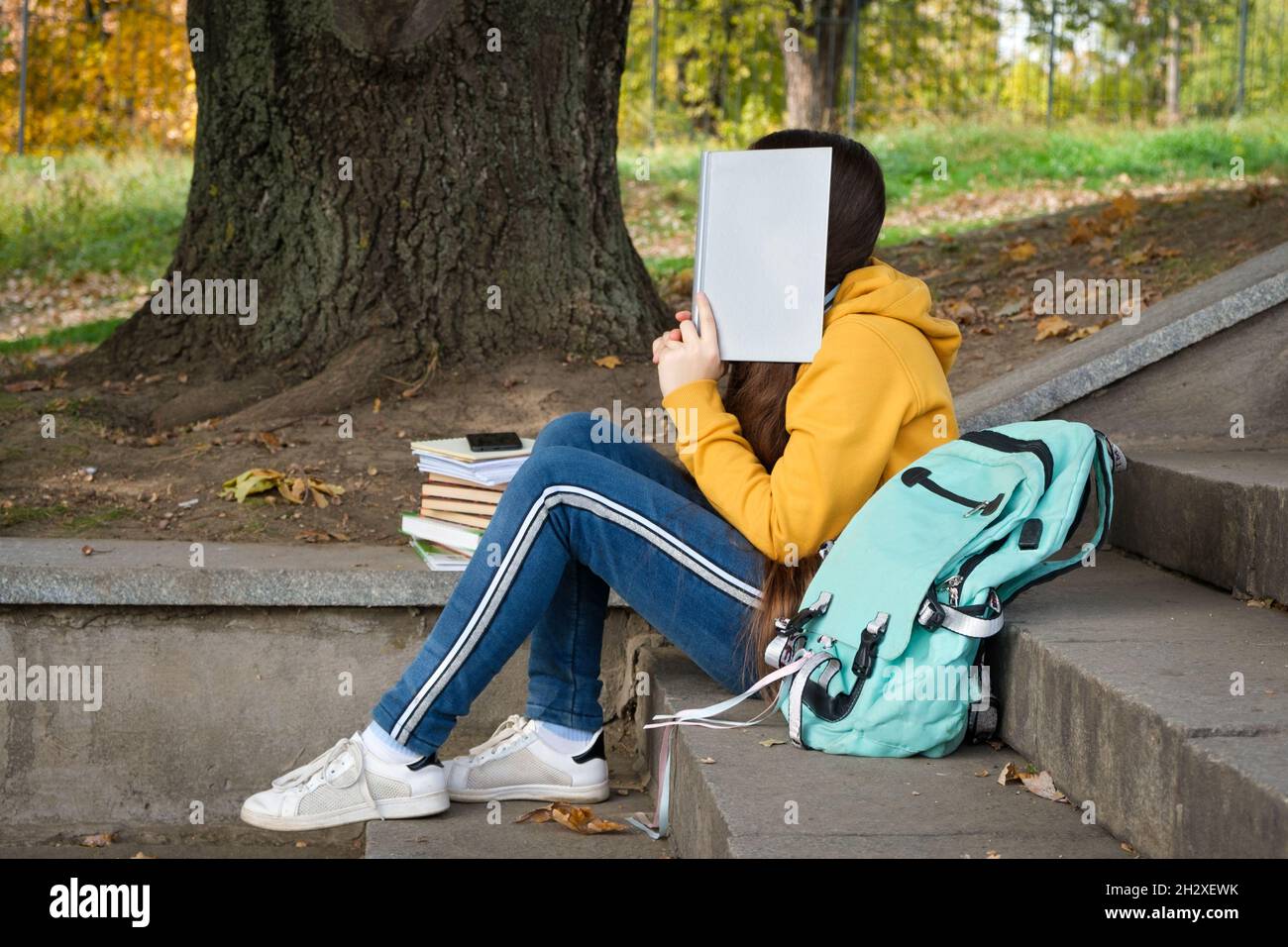 Tired teenage girl reads a book, covers her face with a book sitting on the stairs in the park. Stock Photo
