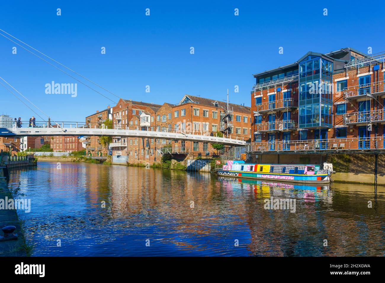 Cable-stayed pedestrianised bridge (footbridge) spanning The River Aire with a colourful fixed barge, Leeds, West Yorkshire, England, UK. Stock Photo