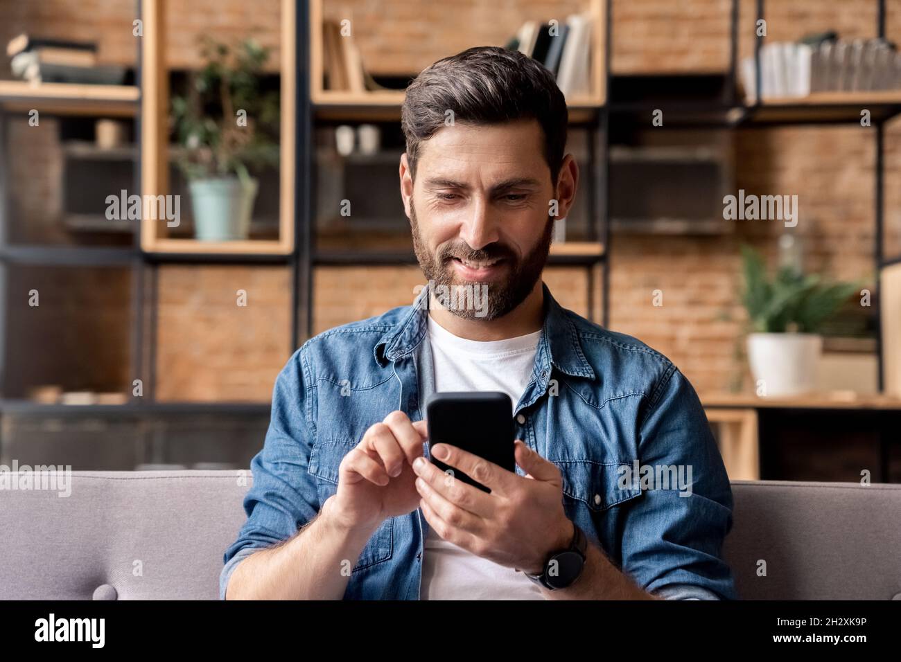 Smiling man using mobile phone scrolling network feed sitting on sofa at  home Stock Photo - Alamy