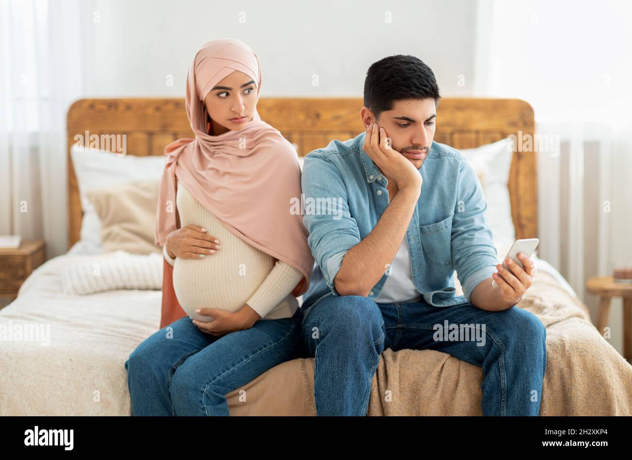 Young pregnant muslim woman offended to her husband busy with smartphone, arab lady in hijab feeling lonely Stock Photo