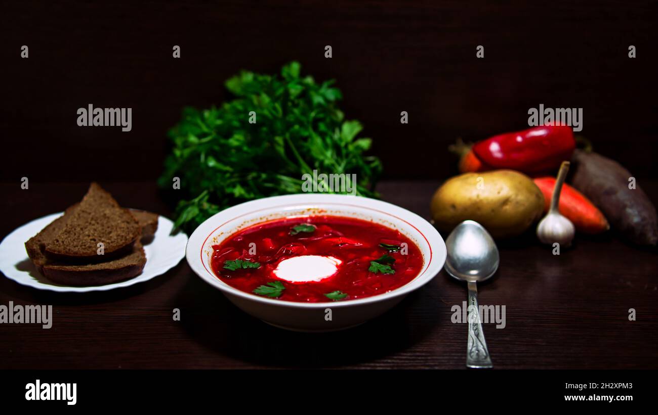 A close-up shot of a plate of red Russian borsch with bread, vegetables, and greens in the background. Stock Photo