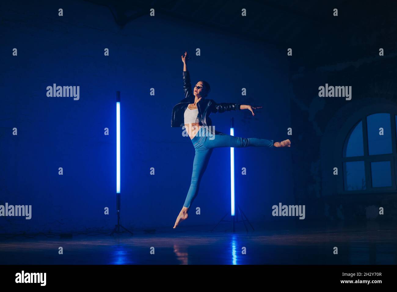 Young woman in casual style - jeans and leather jacket doing ballet in modern studio. Attractive ballerina practices in choreography alone Stock Photo