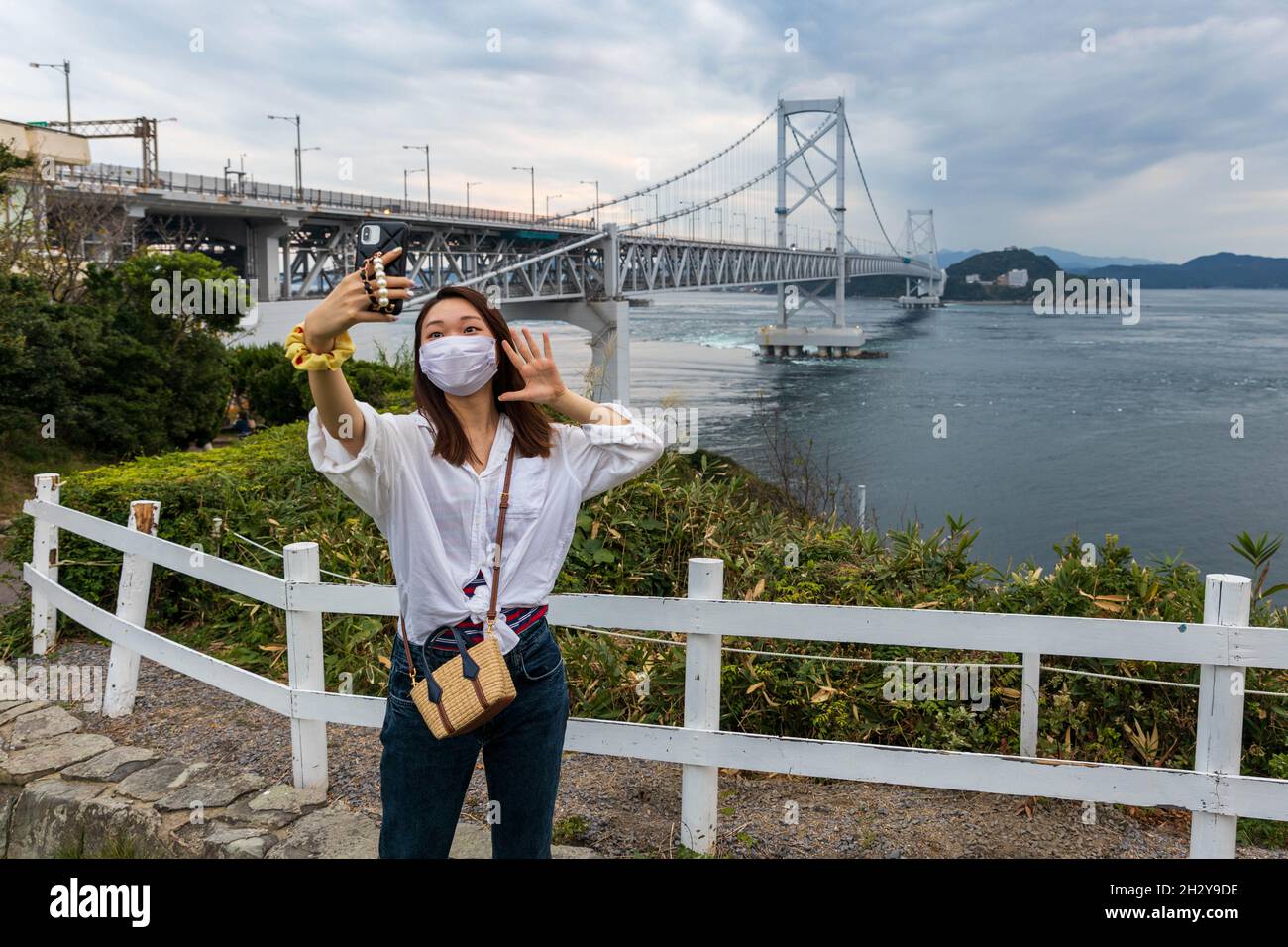 Young woman wearing mask taking selfie on lookout point over suspension bridge and Naruto Straight Stock Photo