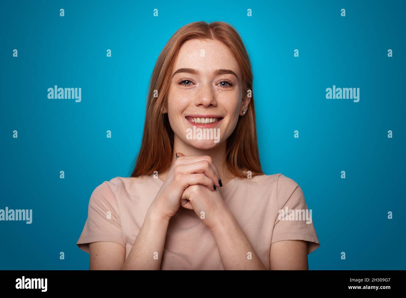 Caucasian young woman posing on a blue background. Smiling and looking at the camera with his hands under his chin. Freckled face Stock Photo