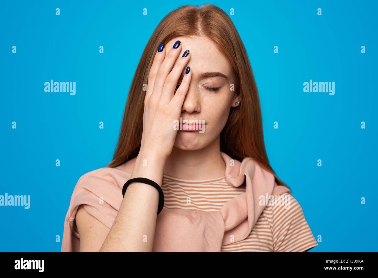 Woman with long red hair covering half of her face with the palm of her right hand. Portrait of a freckled young woman on a blue background Stock Photo