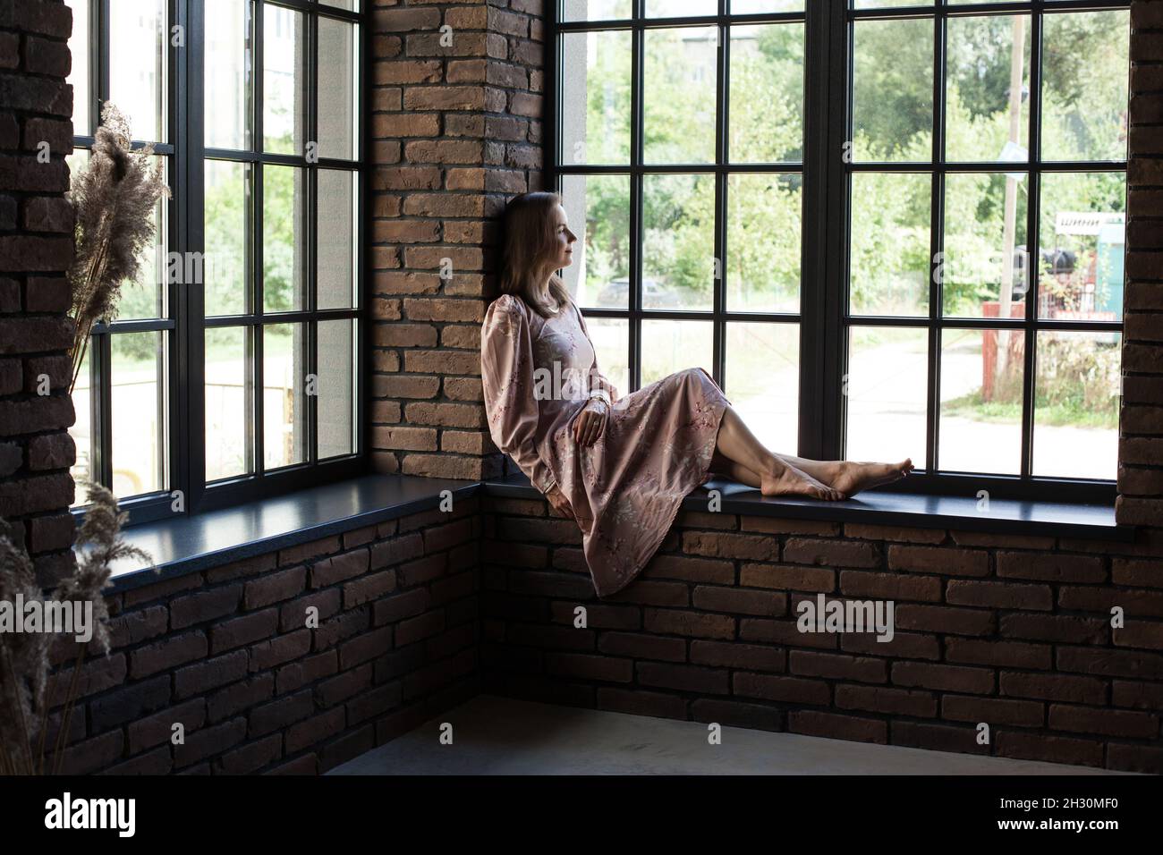 Beautiful young woman in pink dress sits on the windowsill barefoot ...