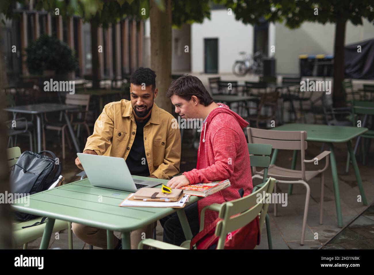 Young man with Down syndrome with his mentoring friend sitting outdoors in cafe using laptop. Stock Photo