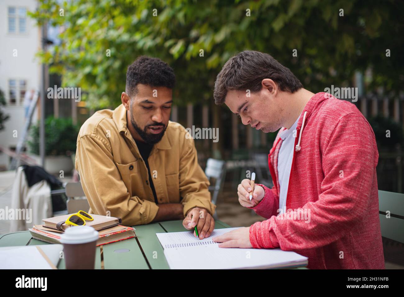 Young man with Down syndrome with his mentoring friend sitting outdoors in cafe and studying. Stock Photo