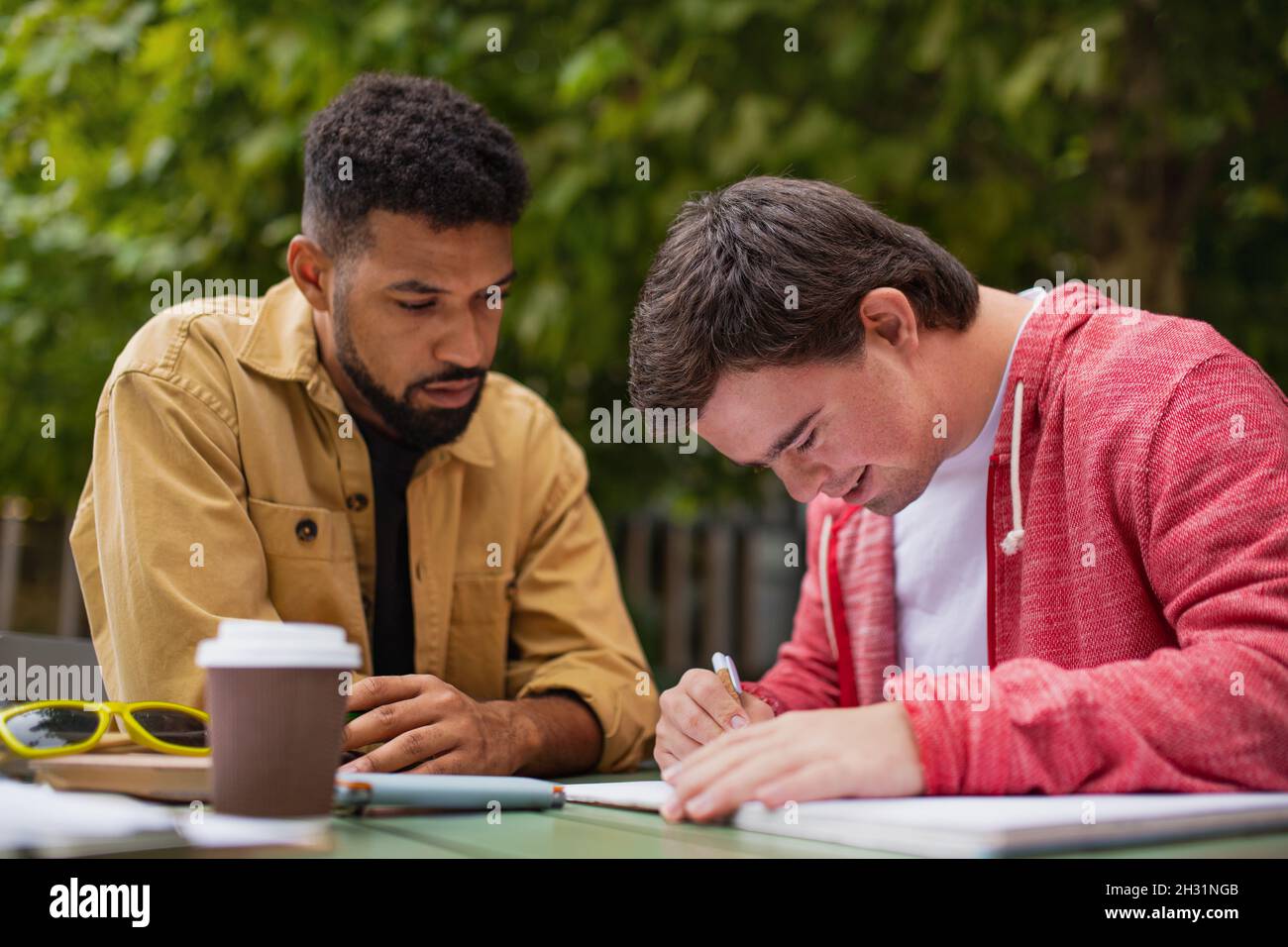 Young man with Down syndrome with his mentoring friend sitting outdoors in cafe and studying. Stock Photo