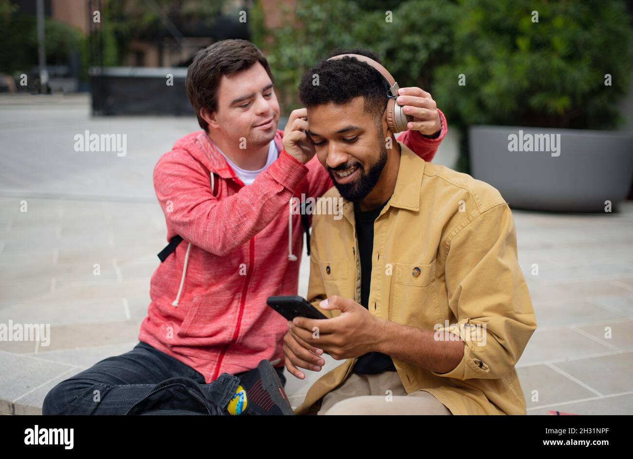 Young man with Down syndrome with his mentoring friend sitting outdoors and listening to music. Stock Photo
