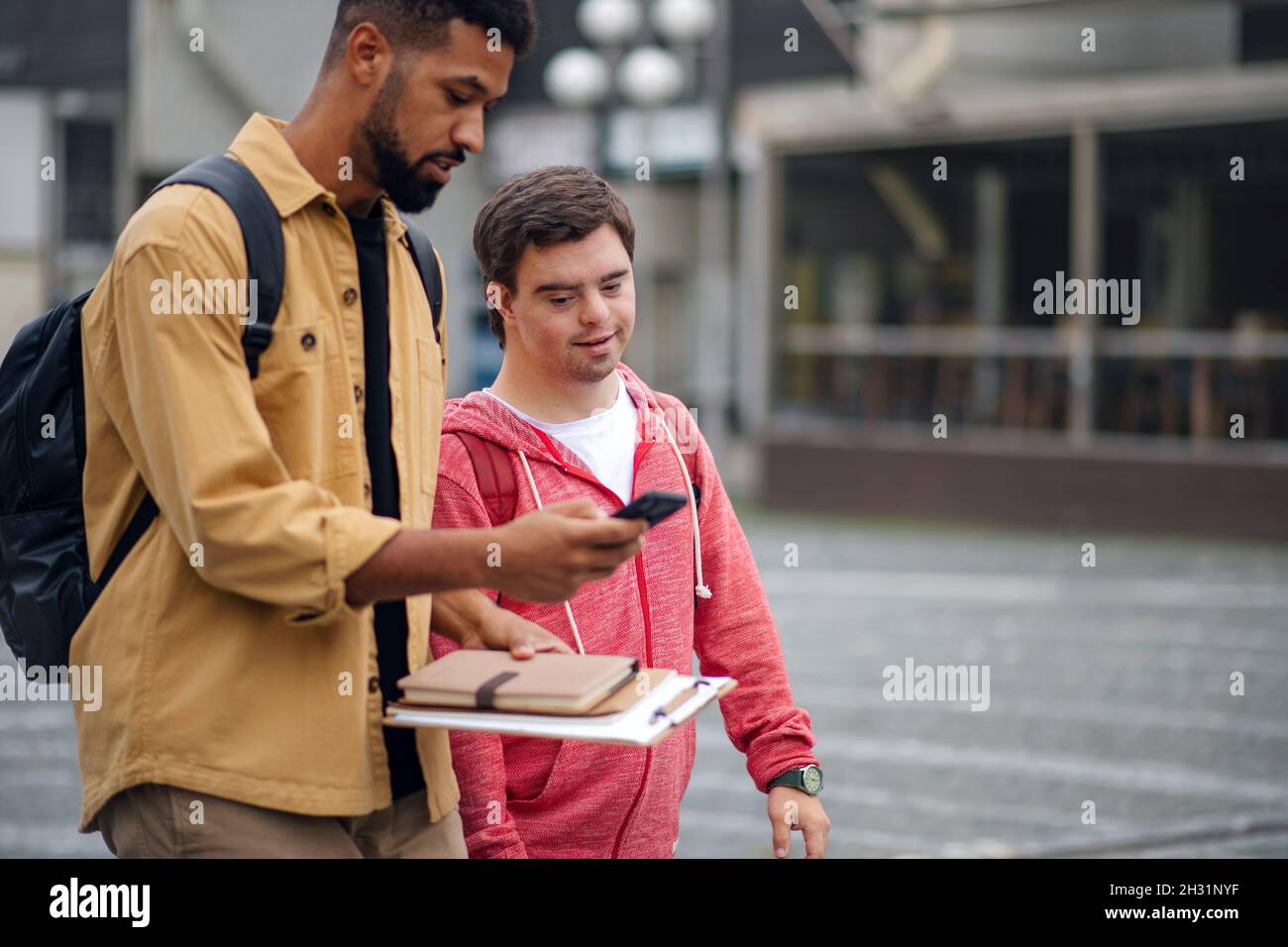Young man with Down syndrome with his mentoring friend with smartphone walking and talking outdoors Stock Photo