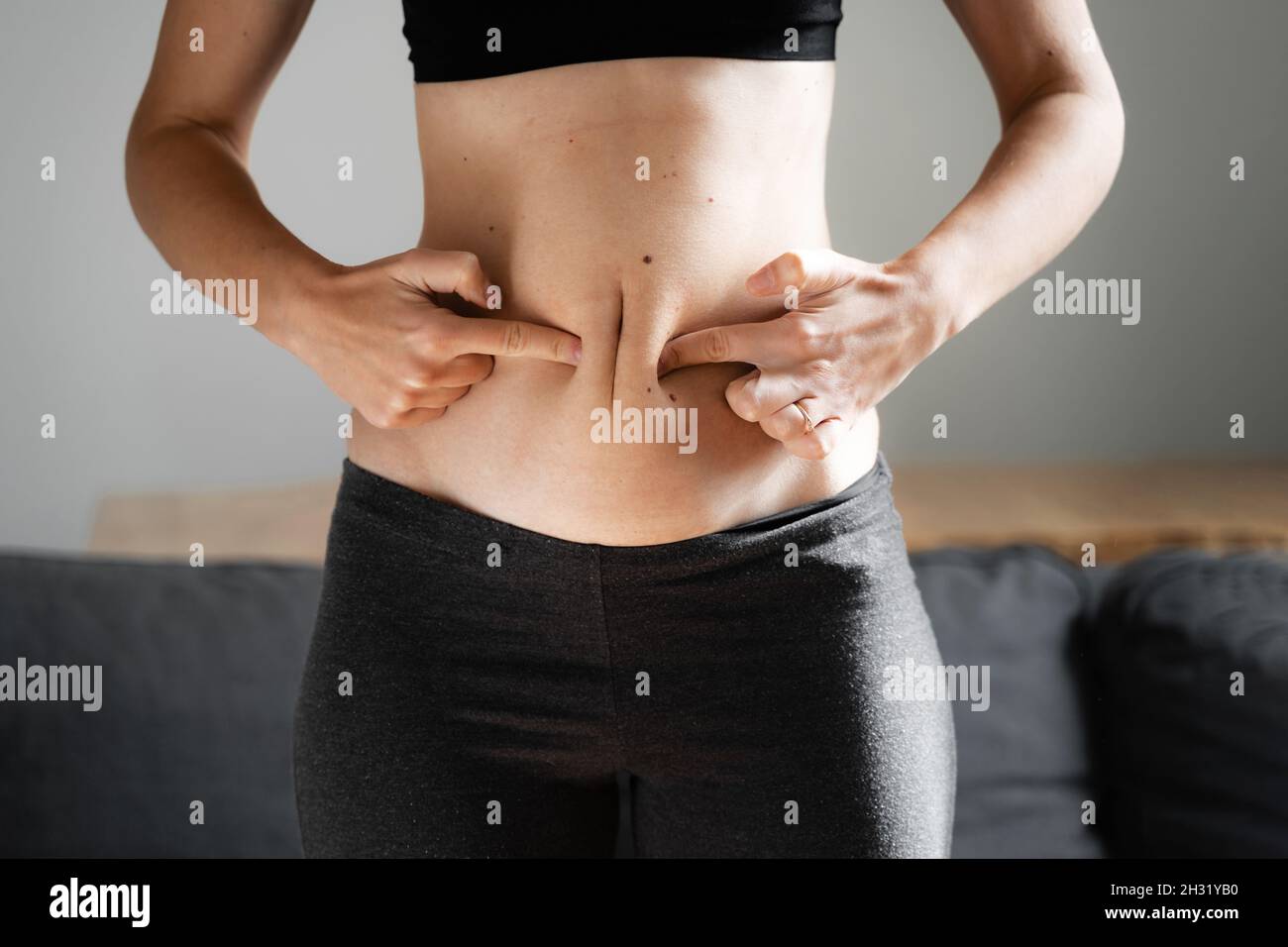 Young woman at home holding belly fat with hands. Body neutrality concept. Accepting the body and weight. Unhealthy carb eating. Stock Photo