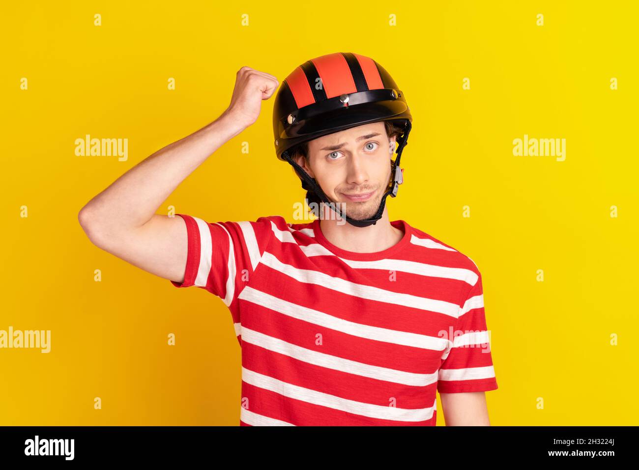 Photo of funny unsure young guy dressed red t-shirt hands arm fist safety hat isolated yellow color background Stock Photo