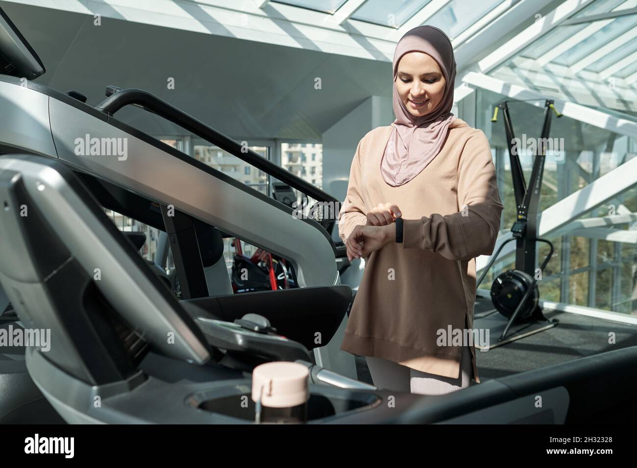 Happy young Muslim woman looking at wristwatch whilestanding by treadmill in large fitness center Stock Photo