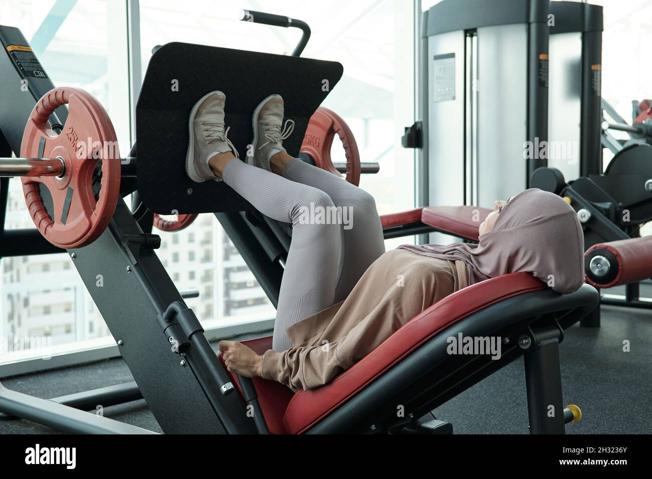 Young Muslim woman pushing part of sports equipment while doing difficult exercise in fitness center Stock Photo