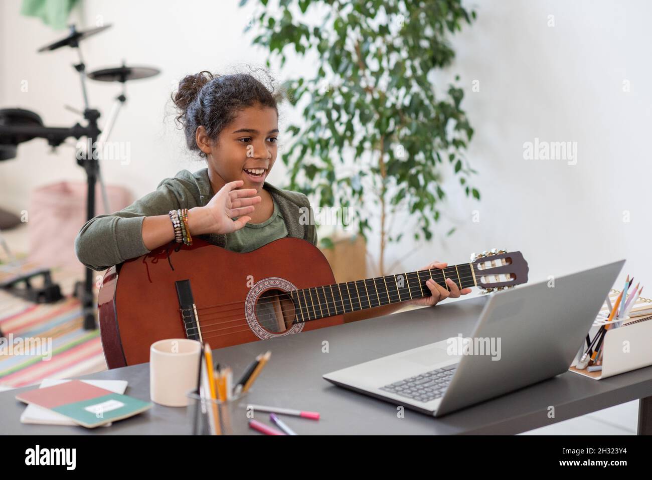 Happy preteen schoolgirl with guitar waving hand to her teacher on laptop screen during online lesson of music Stock Photo