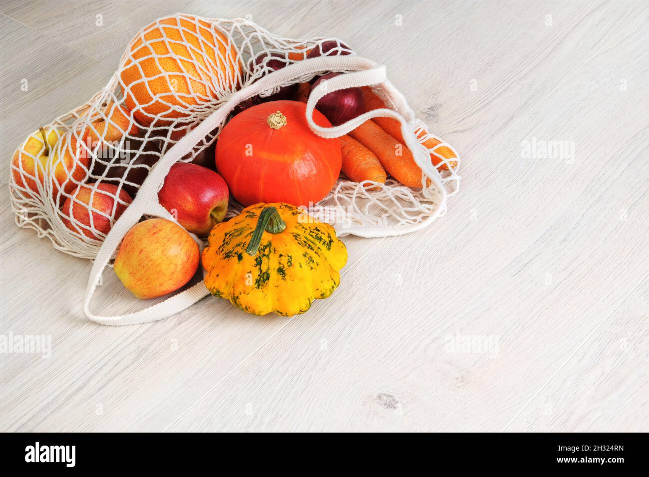 Orange pumpkin, apples, onions, zucchini and carrots in natural reusable mesh bag. Organic vegetables coming out from eco shopping bag. White wooden b Stock Photo