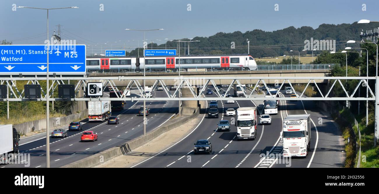 M25 motorway gantry signs railway bridge & Greater Anglia passenger train crossing above road traffic junction 28 for A12 Brentwood Essex England UK Stock Photo
