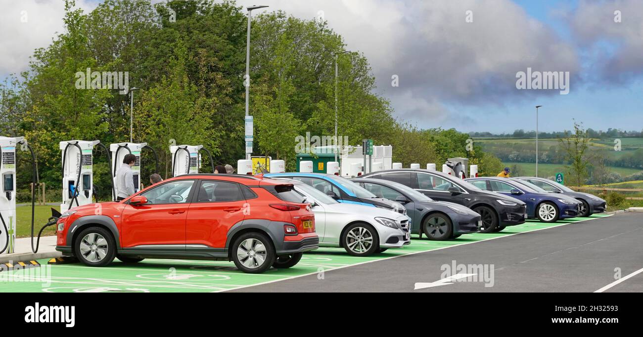 Electric car charging cable to red front bonnet other cars boot tailgate end connected to row of topping up power points green parking bays Rugby UK Stock Photo