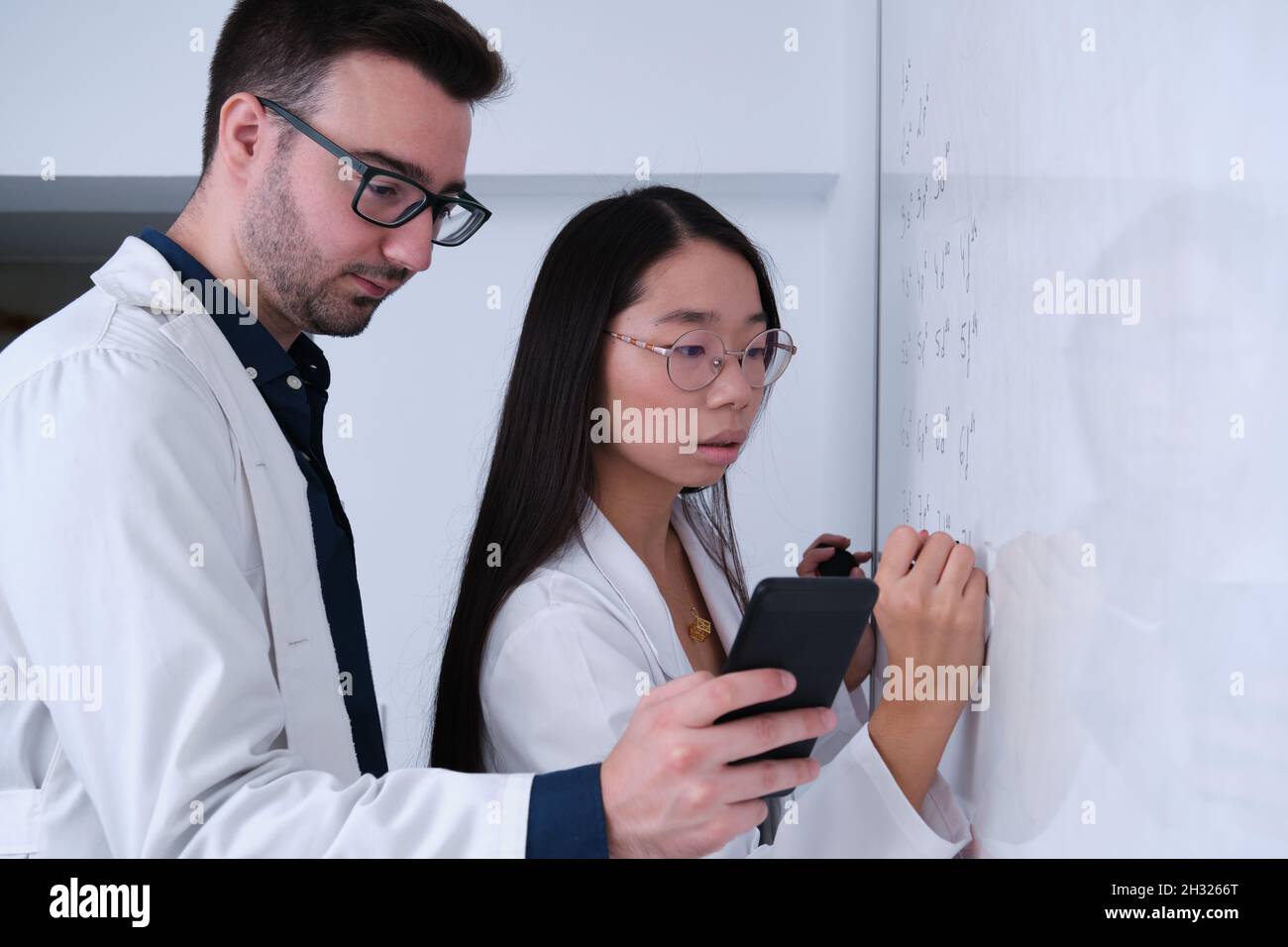 Two researchers in lab coat writing notes on whiteboard. Stock Photo