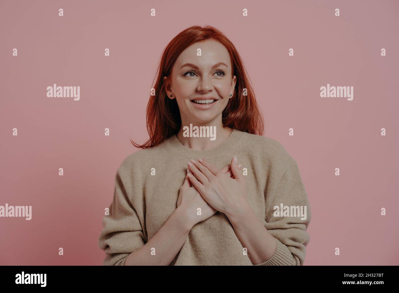 Young happy red-haired woman putting hands on chest while standing isolated on pink background Stock Photo