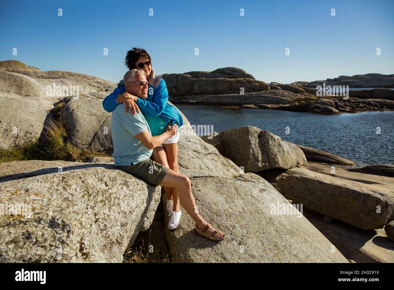 Loving mature couple traveling, sitting on the rock, exploring. Real man and woman hugging, kissing, Happily smiling. Scandinavian landscape, Norway Stock Photo