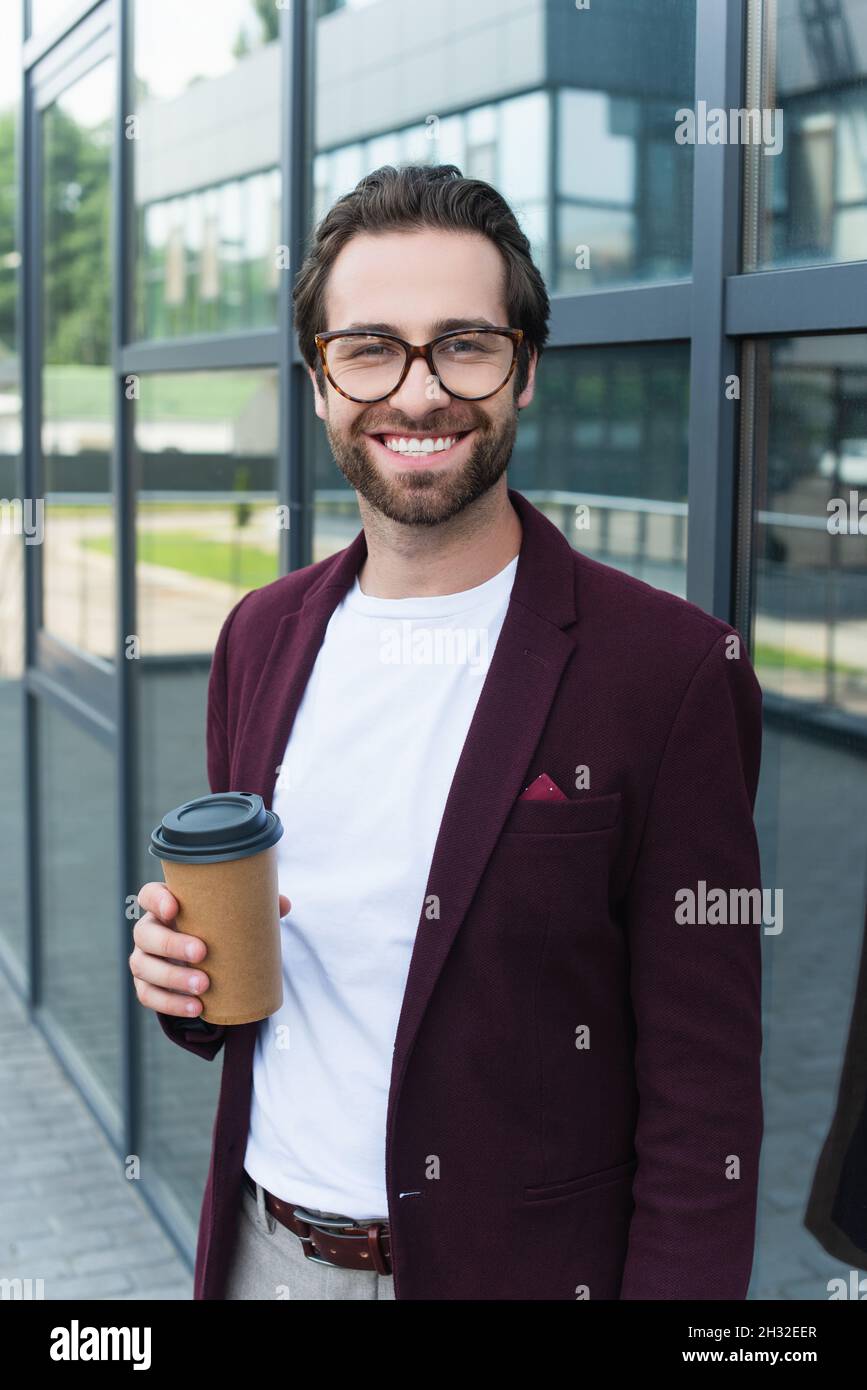 Smiling businessman with coffee to go smiling at camera near building ...
