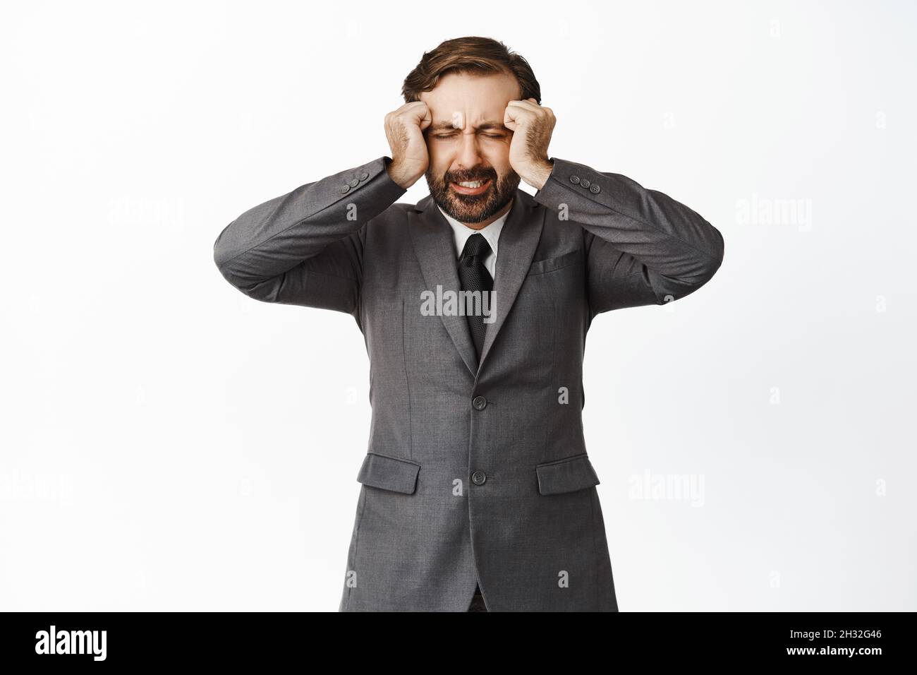 Distressed and tensed corporate man holding hands on head and grimacing, painful headache, frustrated about losing, standing against white background Stock Photo