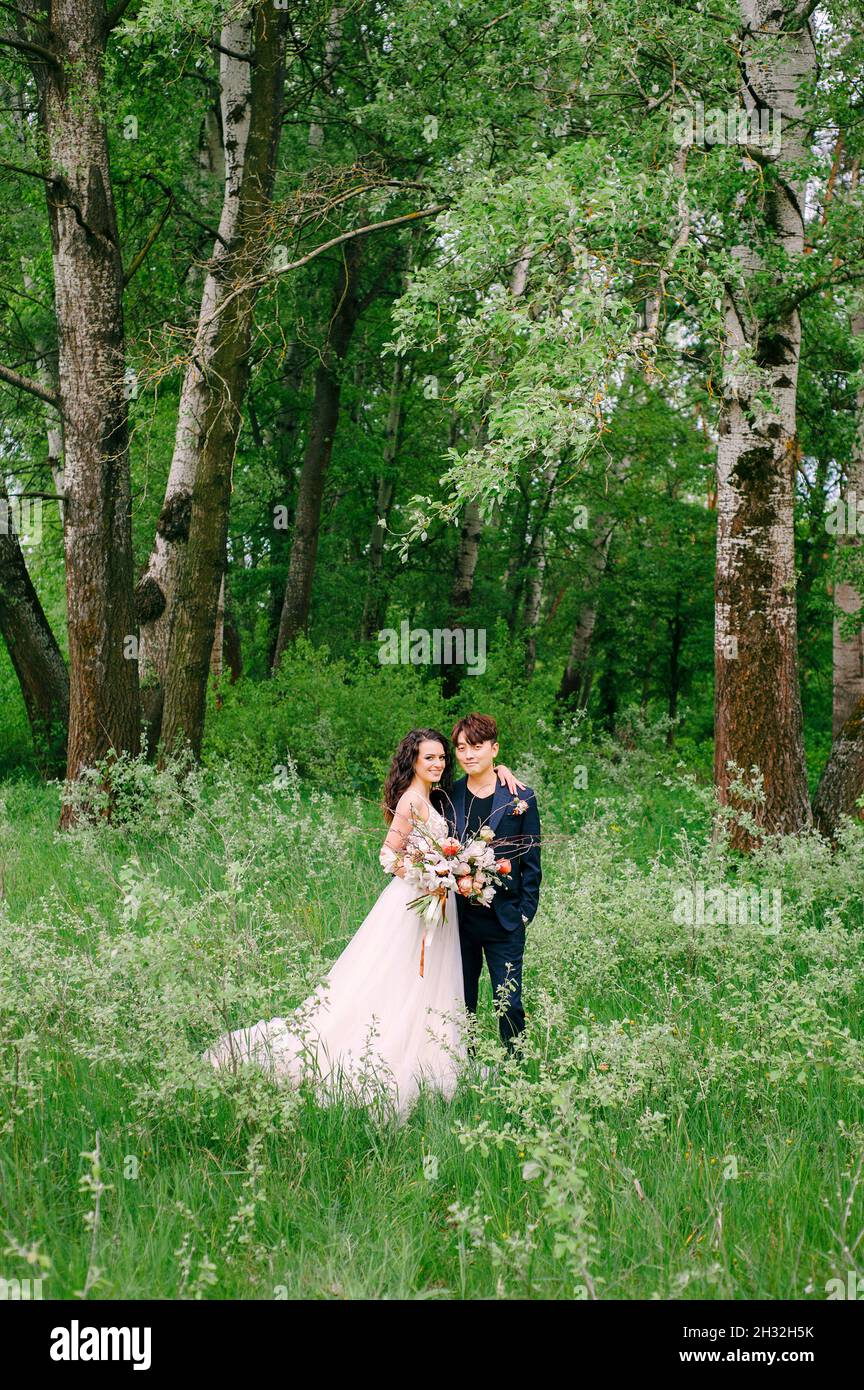 Wedding day. Young happy newlyweds in fresh green forest. Portrait of bride and groom. Spring wedding. International couple. Outdoor wedding ceremony. Stock Photo