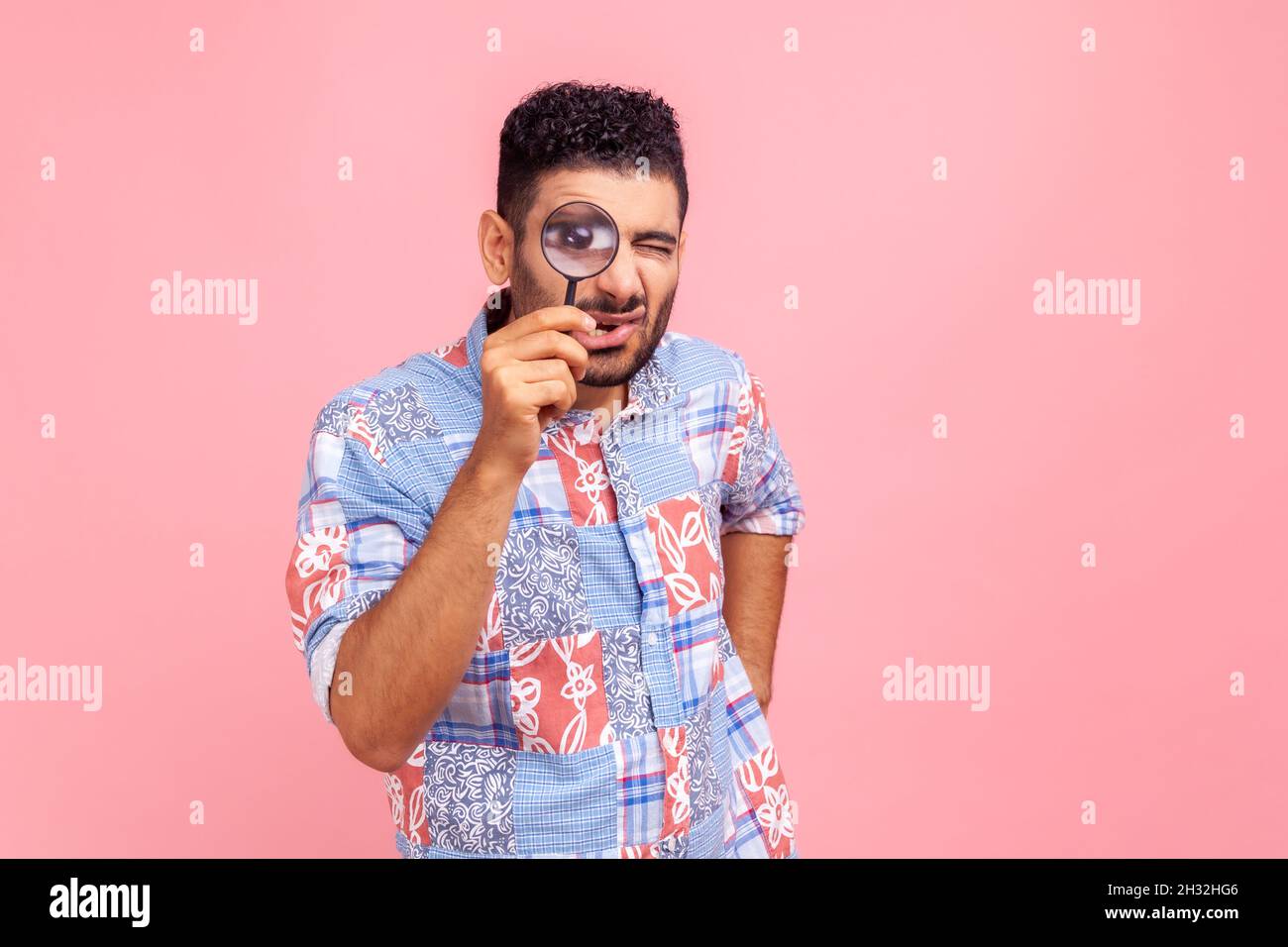Bearded man in blue casual style shirt standing, holding magnifying glass and looking at camera with big zoom eye, having funny facial expression. Indoor studio shot isolated on pink background. Stock Photo