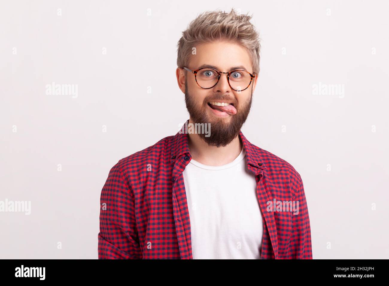 Portrait of funny crazy handsome bearded man in eyeglasses and plaid shirt fooling around and showing tongue, optimism. Indoor studio shot isolated on gray background Stock Photo