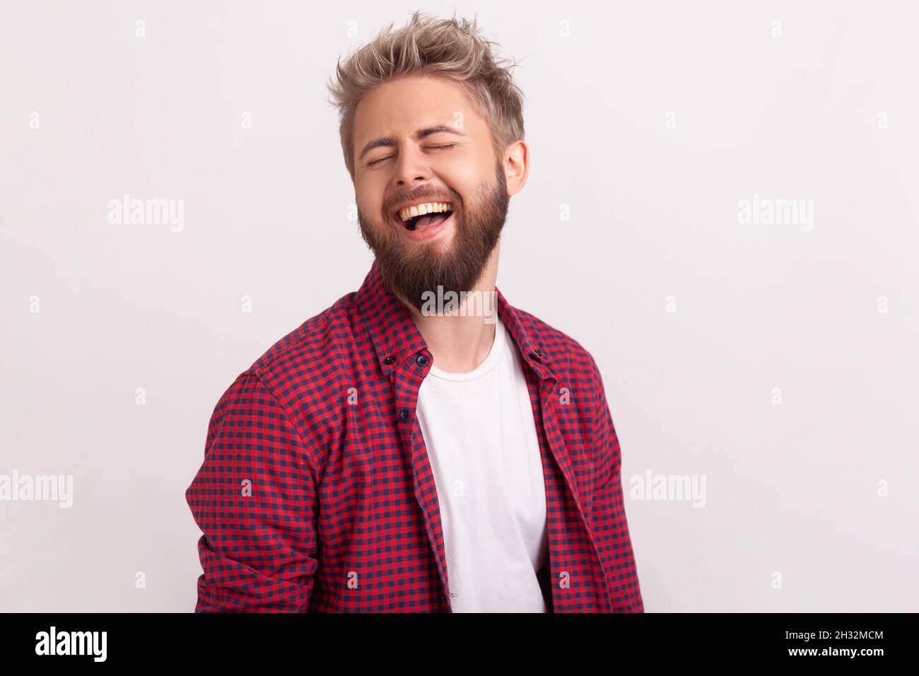 Portrait of funny handsome bearded young man in plaid shirt standing with closed eyes and laughing. Indoor studio shot isolated on gray background Stock Photo