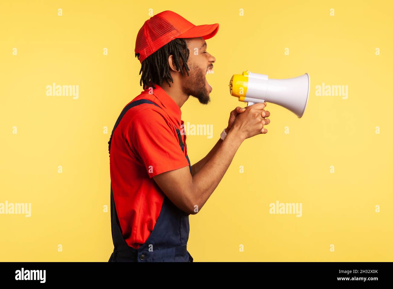 Side view portrait of aggressive worker wearing red T-shirt, cap and blue overalls holding megaphone and screaming loud, protesting. Indoor studio shot isolated on yellow background. Stock Photo