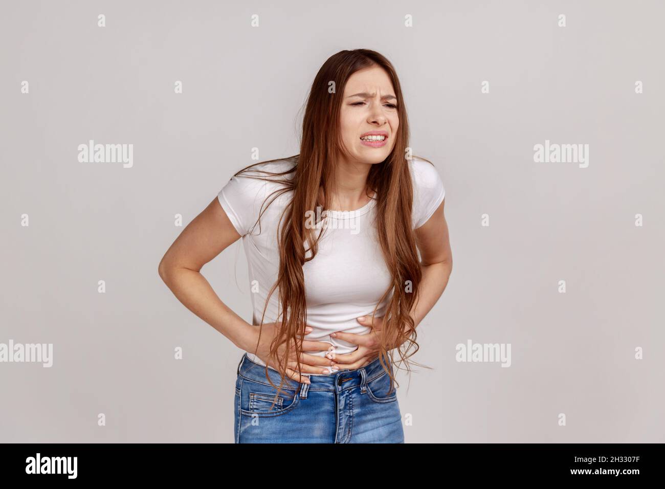 Portrait of unhealthy distressed woman clutching her belly, grimacing from acute abdominal pain, gastritis or constipation, wearing white T-shirt. Indoor studio shot isolated on gray background. Stock Photo