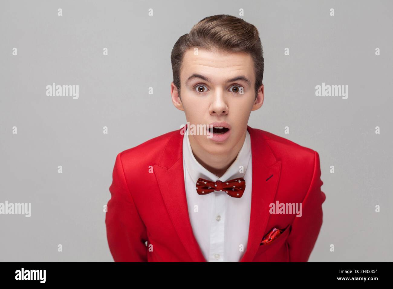 Portrait of handsome gentleman with stylish hairdo in red tuxedo and bow tie standing with big eyes and open mouth, looking at camera in amazement, shock. studio shot isolated on gray background Stock Photo