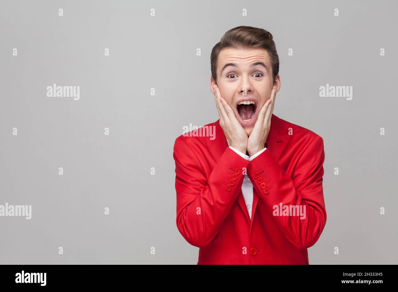 Oh my god. Portrait of extremely amazed gentleman with stylish hairdo in red tuxedo and bow tie holding hands on cheeks and looking at camera with shocked eyes. indoor studio shot, gray background Stock Photo