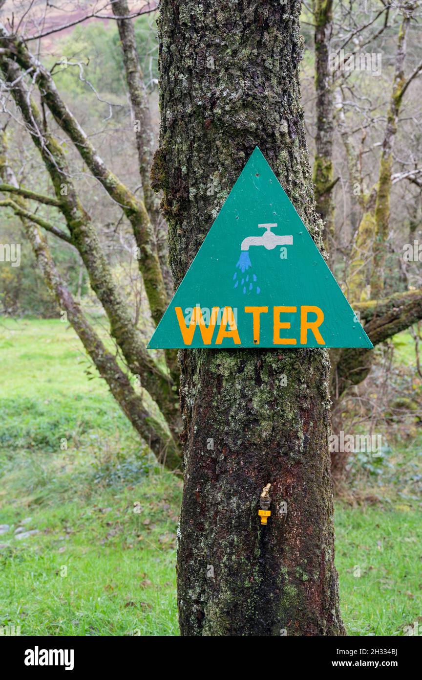 Handpainted triangular sign for water with tap icon, tap underneath is embedded in tree. Red Squirrel campsite, Scotland. Stock Photo