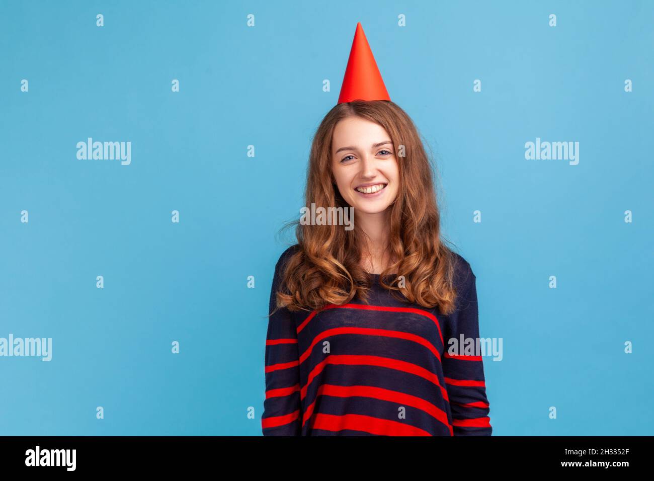 Woman with festive mood wearing striped casual style sweater, standing happy facial expression, celebrating her birthday with party cone on her head. Indoor studio shot isolated on blue background. Stock Photo