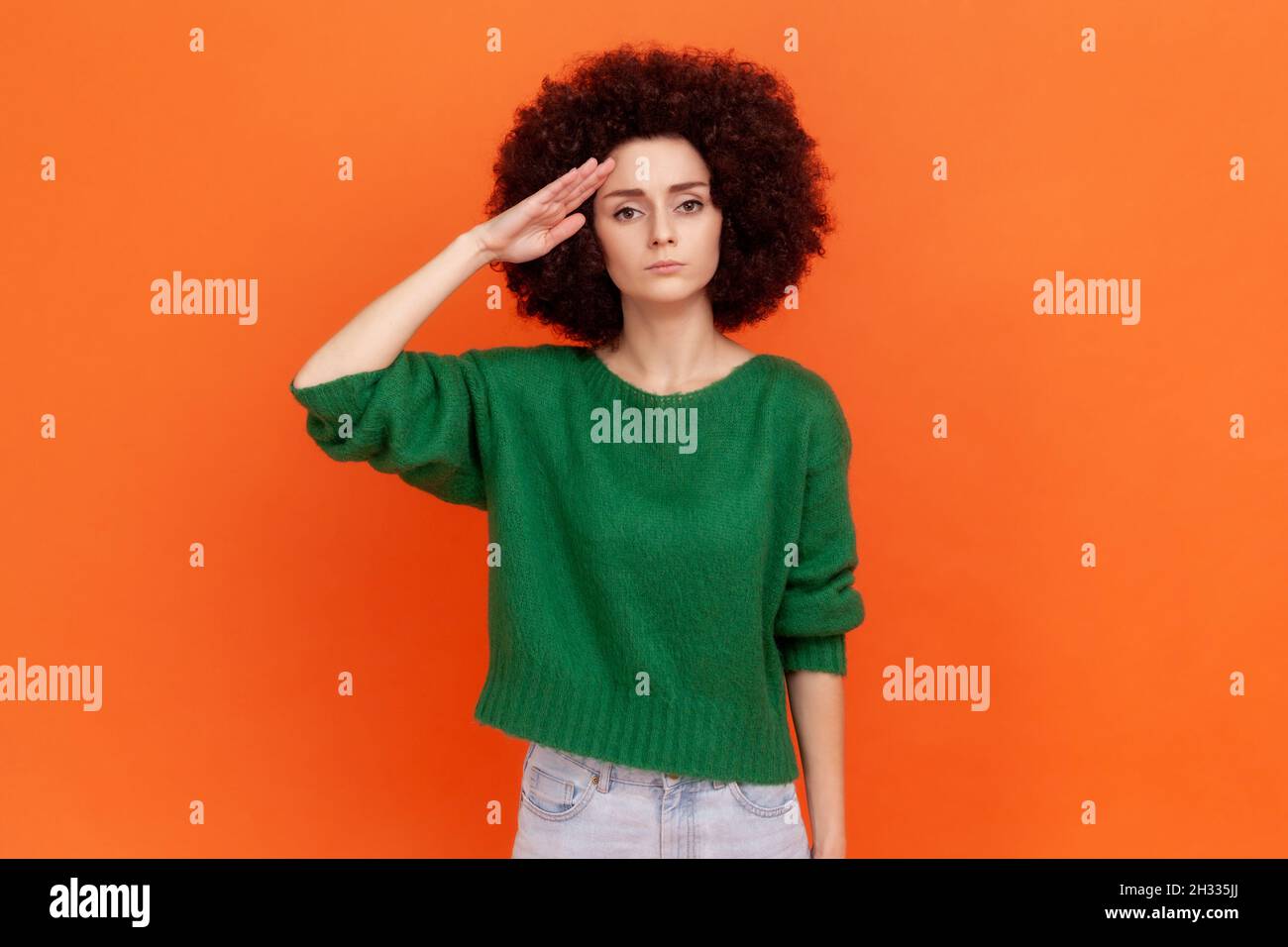 Yes, sir. Strict woman with Afro hairstyle in green sweater saluting with respect and responsibility, patriotic citizen listening order with honor. Indoor studio shot isolated on orange background. Stock Photo